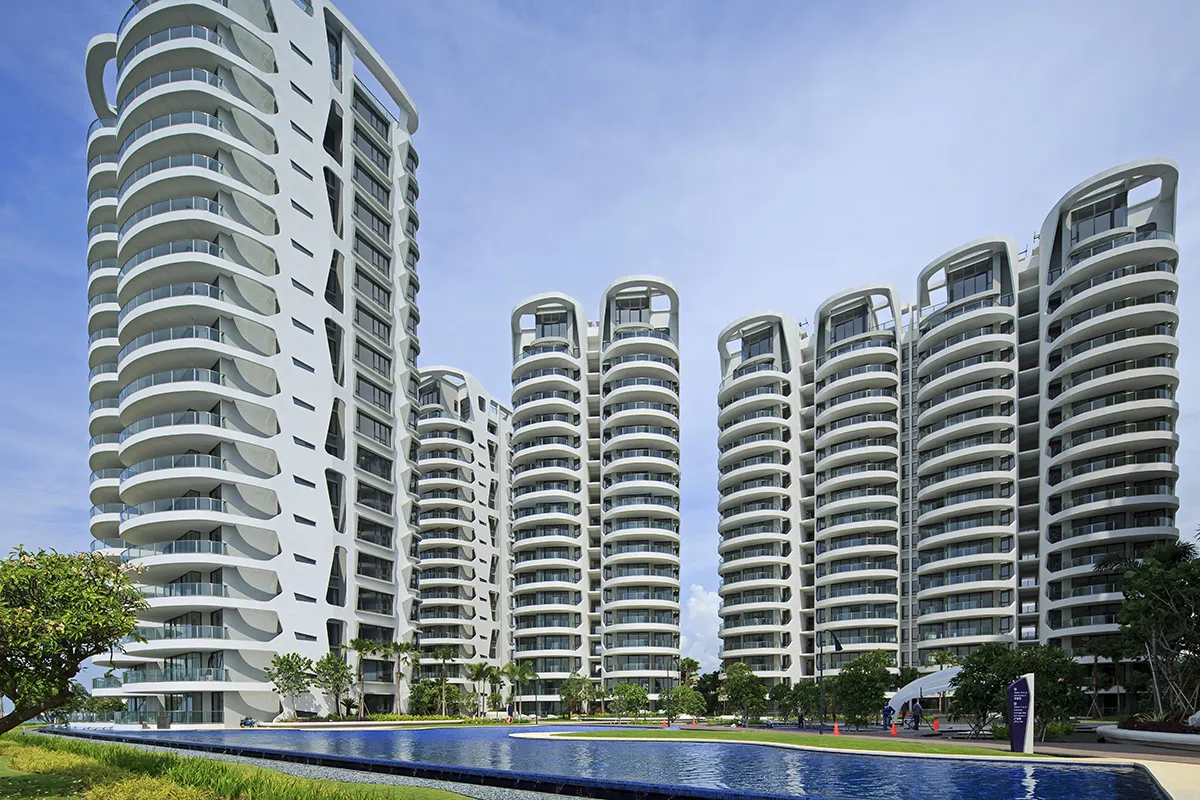 A close-up view of Cape Royale, a luxury waterfront condominium on Sentosa Island, Singapore. The image showcases the modern high-rise towers with sleek curved facades, featuring floor-to-ceiling windows and spacious balconies. In the foreground, a large infinity pool reflects the elegant architecture, surrounded by lush landscaping and tropical greenery. The bright blue sky enhances the striking visual appeal of this premium residential development.