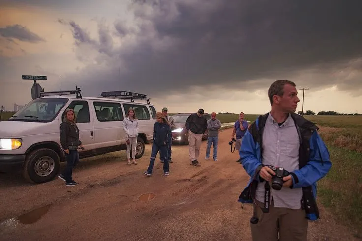 This may contain: a group of people standing in front of a van on a dirt road under a cloudy sky