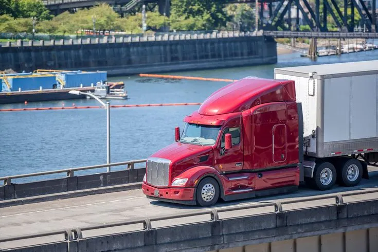This may contain: a red semi truck driving down the road next to a body of water with a bridge in the background