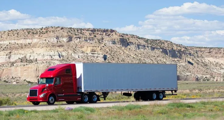 This may contain: a red semi truck driving down the road in front of a large rock mountain range