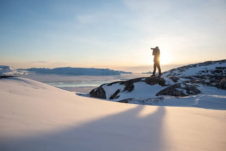 This may contain: a man standing on top of a snow covered mountain with the sun setting behind him