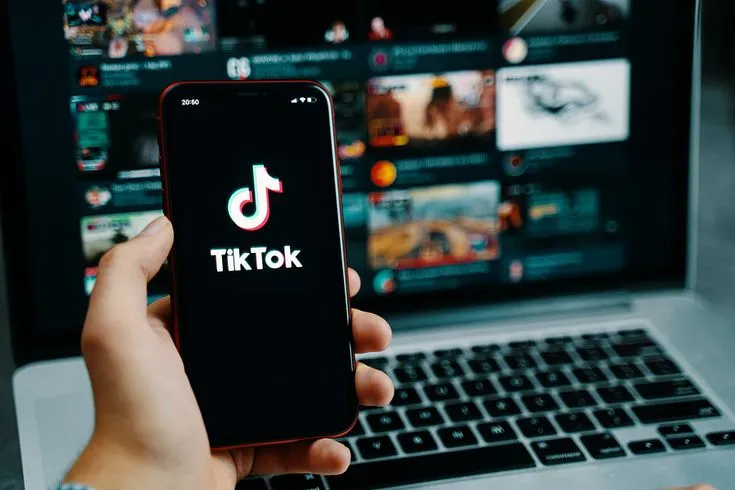 This may contain: a person holding a phone in front of a laptop with the tiktok logo on it
