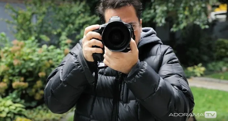 This may contain: a man holding a camera up to his face in front of some bushes and trees
