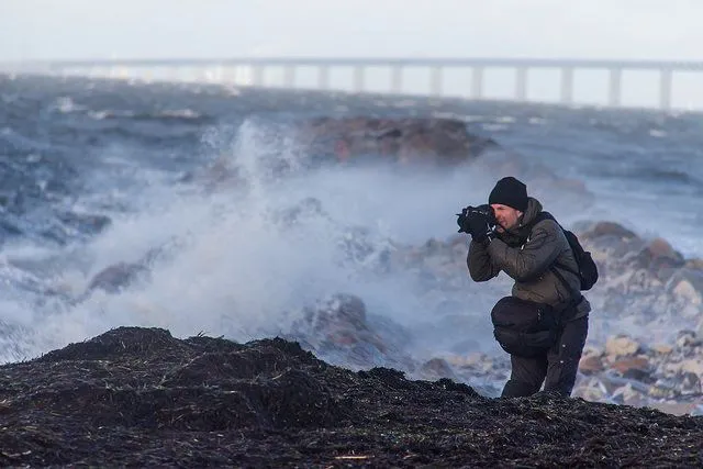 This may contain: a man standing on top of a rocky beach next to the ocean holding a camera