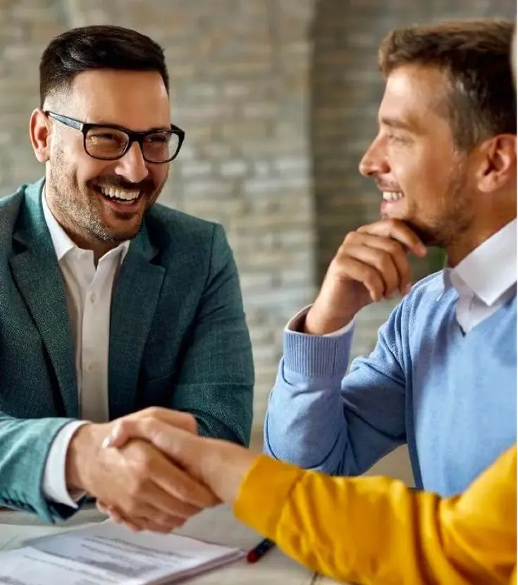 This may contain: two men shaking hands while sitting at a table in front of a laptop computer and papers