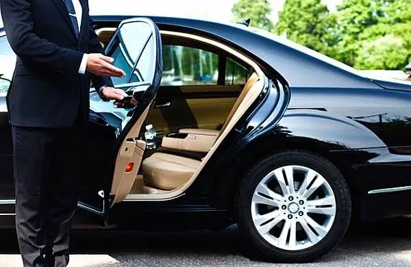 This may contain: a man in a suit standing next to a black car with its door open and his hand out