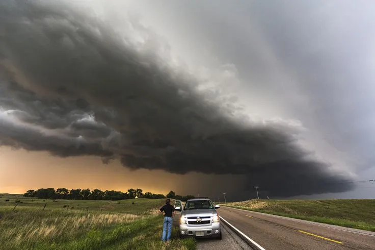 This may contain: a man standing next to a car on the side of a road under a storm cloud