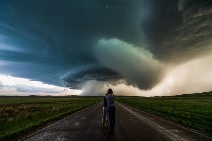 This may contain: a woman standing on the side of a road under a large storm in the distance