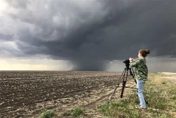 This may contain: a woman standing on top of a grass covered field next to a large storm cloud