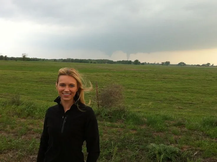 This may contain: a woman standing in the middle of a field with storm clouds above her and trees behind her