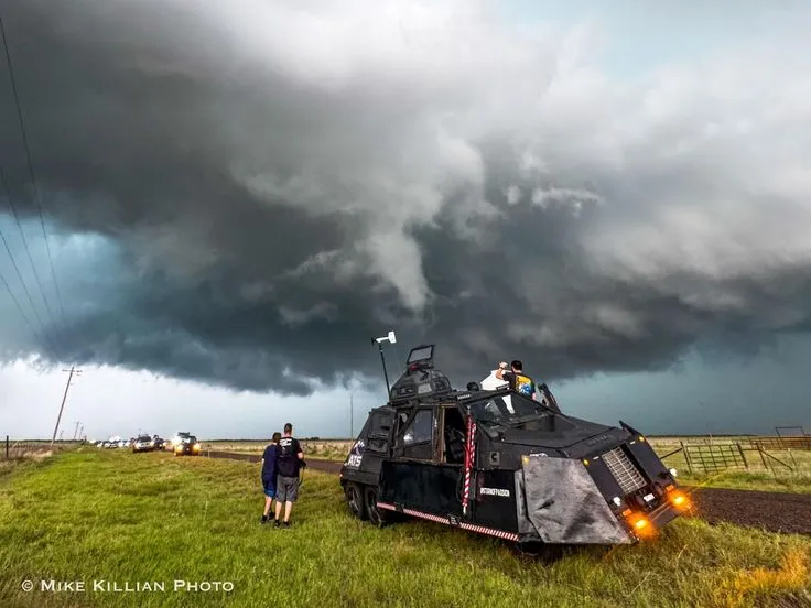 This may contain: two people standing in front of an armored vehicle under a cloudy sky with dark clouds