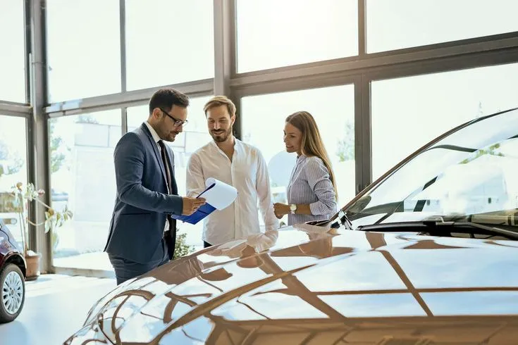 This may contain: two men and a woman standing in front of a car talking to each other while holding papers