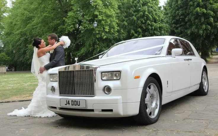 This may contain: a bride and groom kissing in front of a white rolls royce wedding car on the street