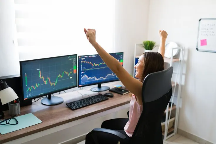 This may contain: a woman raising her arm in the air while sitting at a desk with two computer monitors