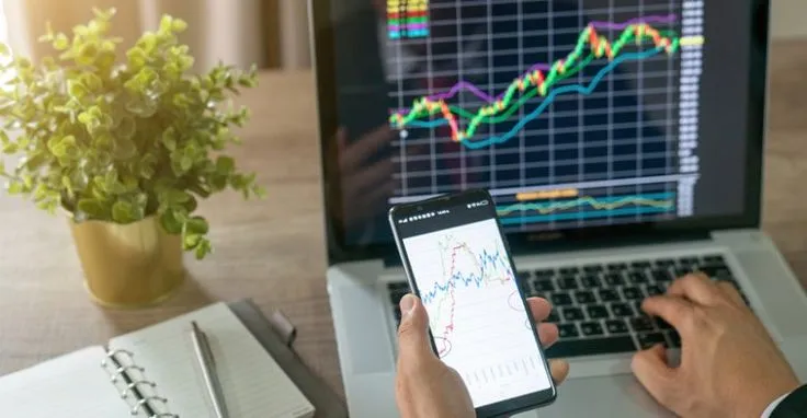 This may contain: a man is using his cell phone and laptop to check the stock market on their computer screen