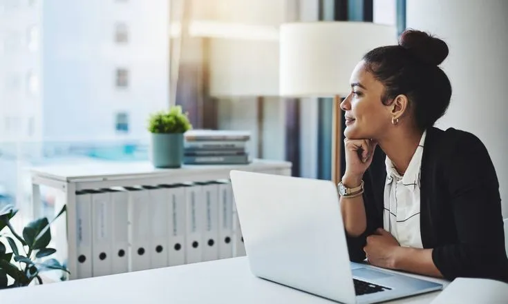 This may contain: a woman sitting in front of a laptop computer on top of a white desk next to a window