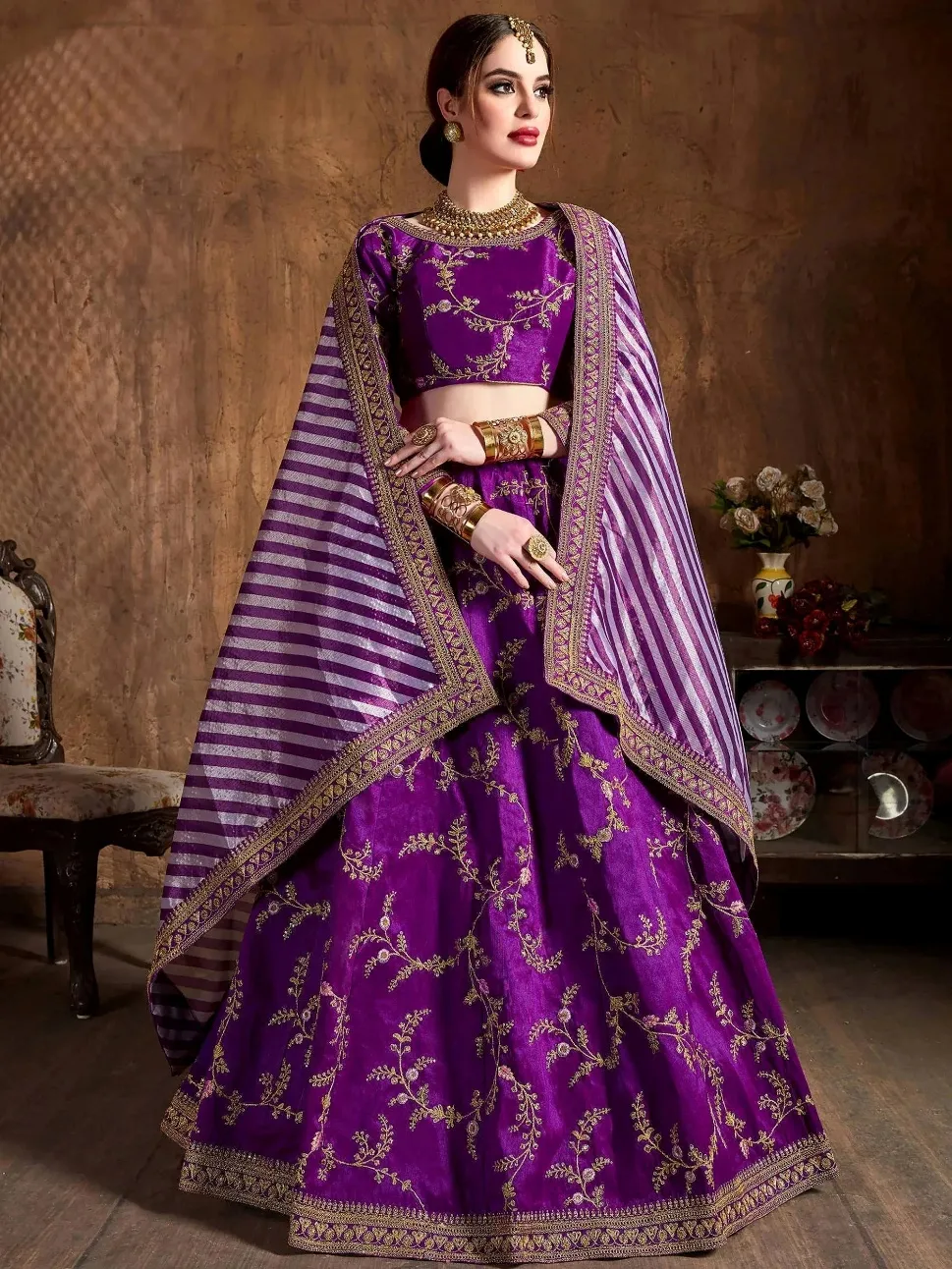 Woman wearing a purple designer lehenga choli with gold embroidery and a striped dupatta