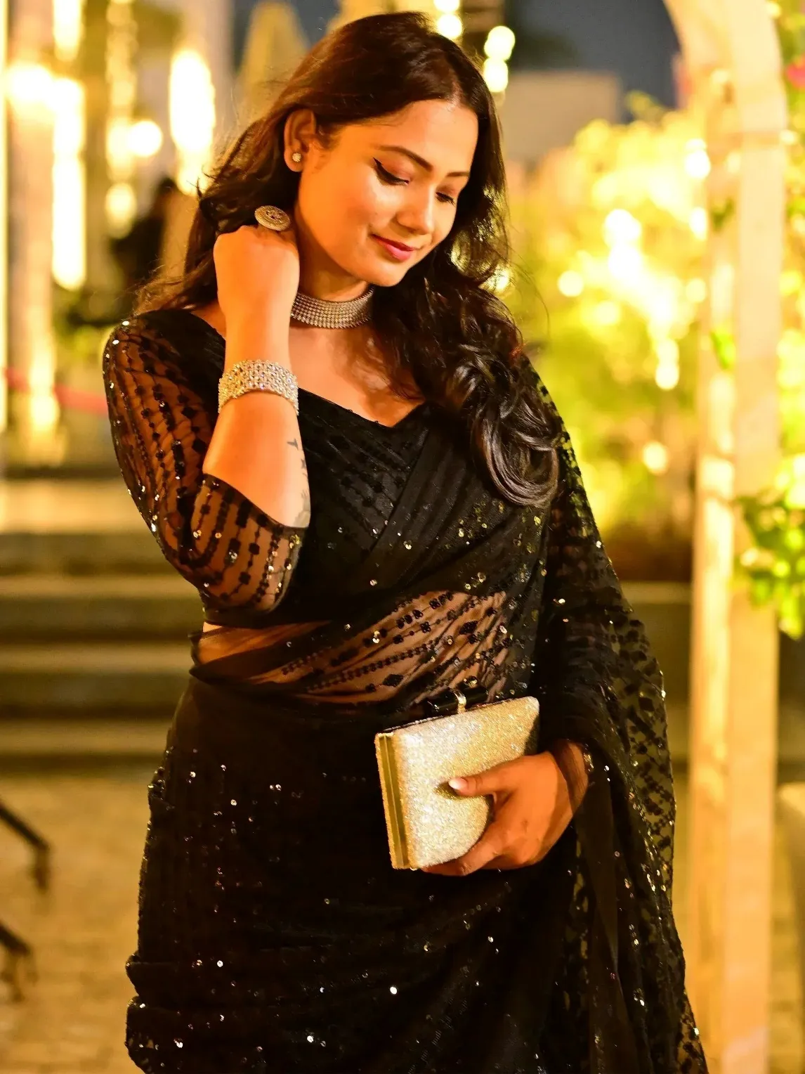 Elegant woman in black sequin saree with sheer blouse, holding gold clutch, perfect for Indian ethnic events.